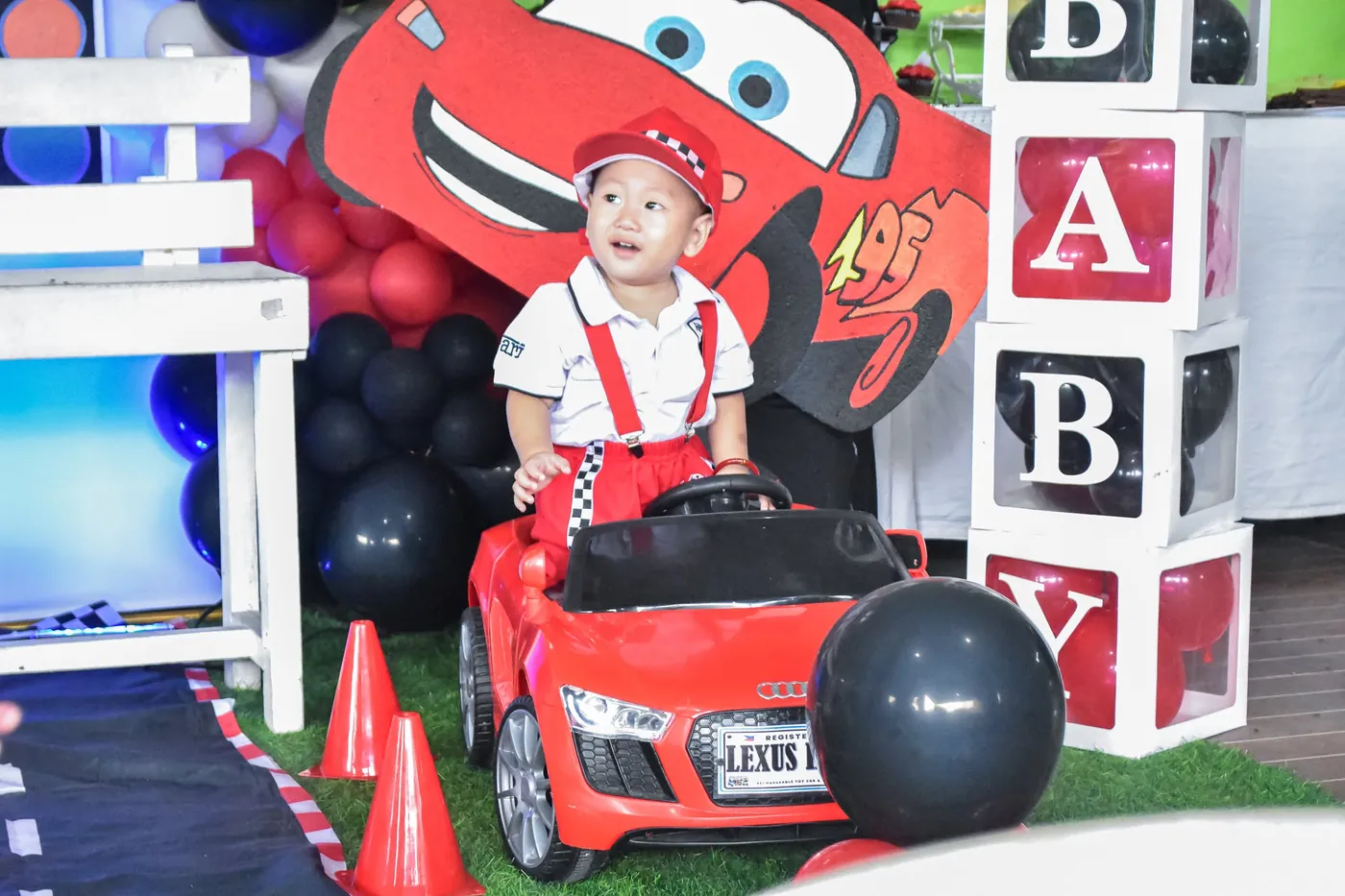 Toddler in a red racing outfit on a toy red car in front of a Cars-themed balloon backdrop at a birthday celebration
