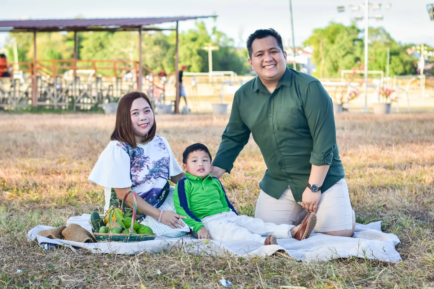Family of three on a picnic blanket with a basket of fruit in a park