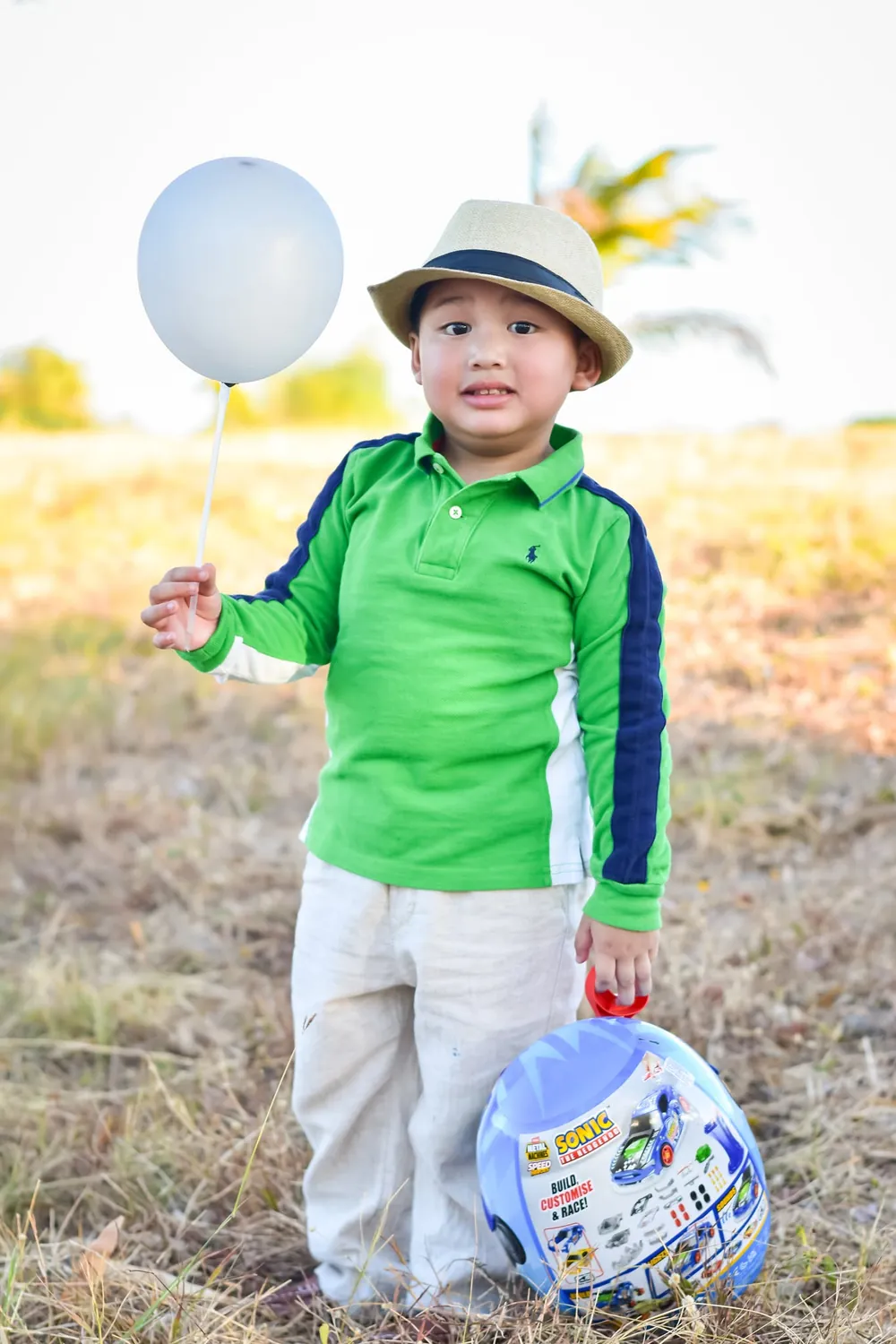 Toddler in a green polo and straw hat holding a white balloon, outdoor portrait